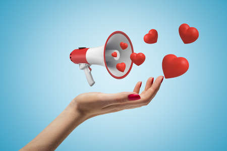 Side closeup of woman's hand facing up and levitating small megaphone that emits cute red hearts, on light blue background. Love and relationship. Express your feelings. Send love message.の写真素材
