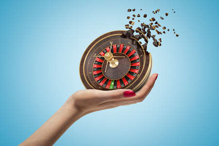 Female hand holding casino roulette shattering into small pieces on blue background. Gambling and betting. Risk and reward. Digital art.の写真素材