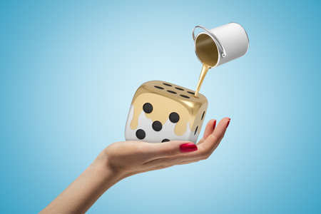 Closeup of womans hand facing up holding white die cube and can of gold paint in air poring paint onto die on light-blue background.の写真素材