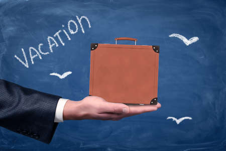 Side view of man's hand facing up and holding brown travel case against blackboard with chalk birds and title 'Vacation'. Travel vacation. Travel agencies. Baggage insurance.の写真素材