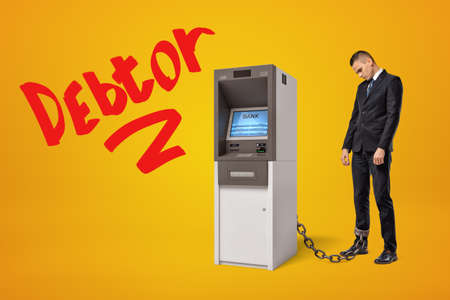 Sad young businessman standing in half-turn chained to ATM on amber background with big red title Debtor.の写真素材