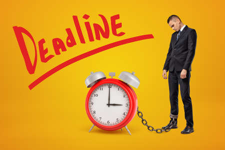 Sad, tired businessman standing in half-turn chained to huge red alarm clock on amber background with big red title Deadline.の写真素材