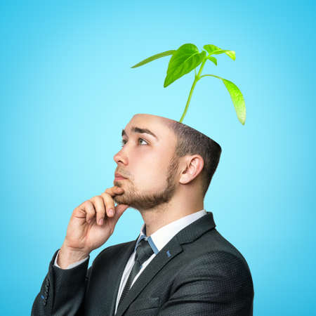 Crop image of young man in suit, looking up and thinking with hand at chin, top of head cut off and green sprout growing from it.の写真素材