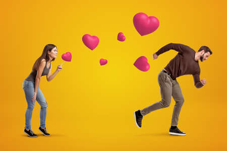 Side view of young girl following with her eyes young man whos running away being chased by cute valentine hearts that float in air on amber background.の写真素材
