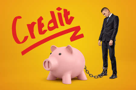 Sad businessman chained to pink piggy bank with Credit sign above on yellow background.の写真素材