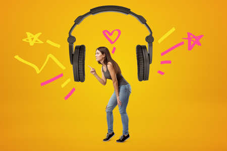 Side view of young beautiful girl standing between earcups of huge black earphones suspended in air playing loud music on amber background.の写真素材