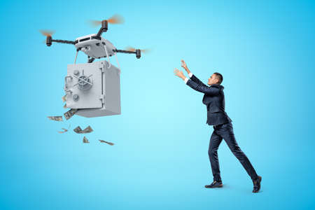 Businessman raising his hands to big quadcopter carrying money safe with open door and money falling out on blue background.の写真素材