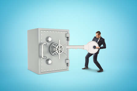 Tiny businessman holding silver key next to big metal bank safe on blue backgroundの写真素材