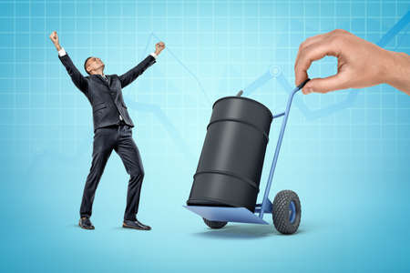 Very happy businessman standing near big black oil barrel on hand truck held by huge hand on blue background. Oil industry. Successful business. Fuel transportation.の写真素材