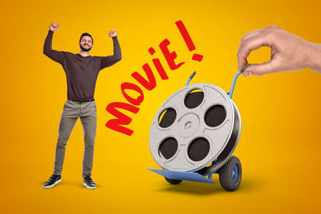 Happy man in casual clothes and hand holding hand truck with film reel and Movie sign on yellow background.の写真素材