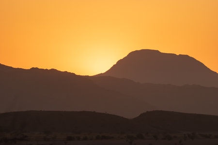 A warm sunset illuminates the silhouettes of distant mountains. The golden hues create a serene and peaceful desert atmosphere.の写真素材