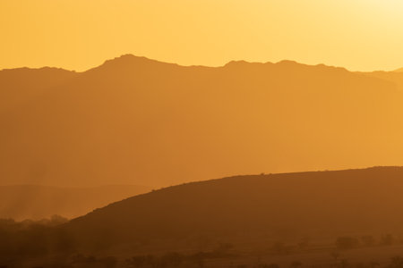 A warm sunset illuminates the silhouettes of distant mountains. The golden hues create a serene and peaceful desert atmosphere.の写真素材