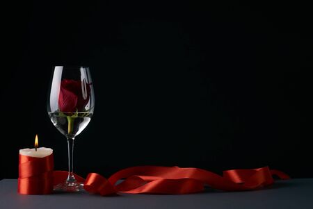Wineglass with rosebud inside, candle and red ribbon on dark background. Love card concept with copy space. Valentine's day themeの写真素材