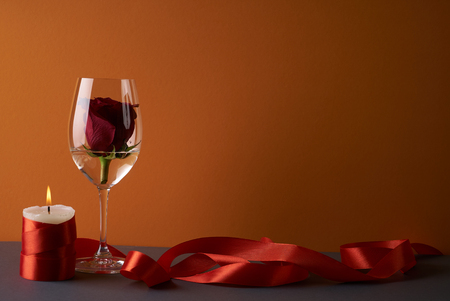 Wineglass with rosebud inside, candle and red ribbon on orange background. Love card concept with copy space. Valentine's day themeの写真素材