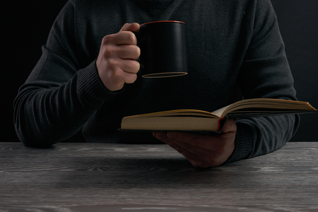 Man in dark sweater sitting by wooden table and reading book while holding mugの写真素材