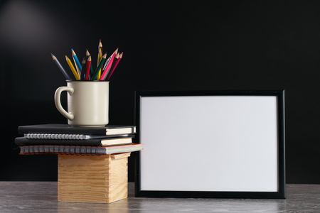 Desk with blank white picture frame and pencils in iron mug placed on books and wooden box. Dark backgroundの写真素材