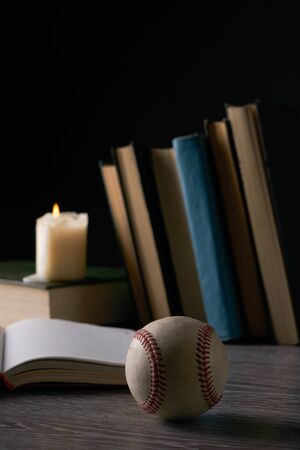 Books, baseball ball and candle on wooden table against black. Education, knowledge conceptの写真素材