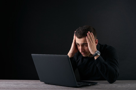 Feeling sick and tired. Frustrated young man holding head in hands while looking to laptop, Dark...