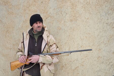 Hunter man dressed in camouflage clothing on the old clay wall. Armed with a rifle.