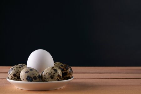 Chicken and quail eggs in dish plate on wooden table against black background. Close up with copy spaceの写真素材