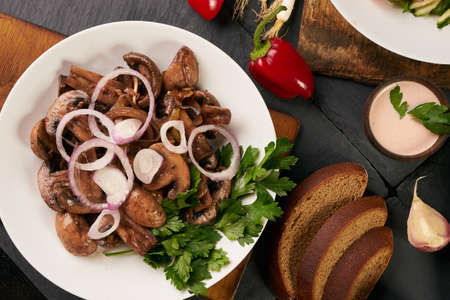 Cooked fried mushrooms on white plate over dark stone background with rye diet bread, low-calorie sauce, fresh red bell pepper and fragrant garlic, top view. Healthy vegetarian food and detox conceptの写真素材