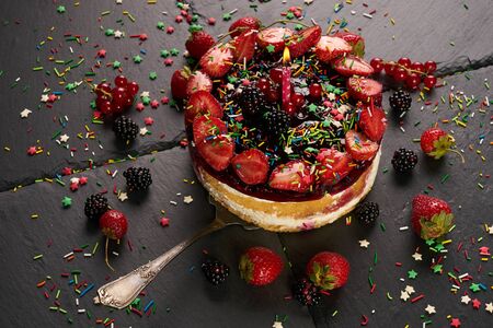 Fresh delicious birthday raspberry cake with fresh strawberries, raspberries, blueberry, currants decorated with holiday candle isolated on dark stone background.の写真素材