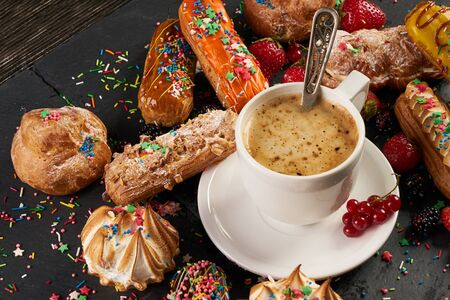 Breakfast cakes French eclairs with white icing, special plum eclairs, mint, blueberries, strawberries and cup of hot coffee drink on a cutting board on a dark black stone backgroundの写真素材