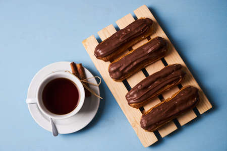 Cup of hot tea with cinnamon steak and french chocolate eclairs on wooden cake board on blue background.の写真素材