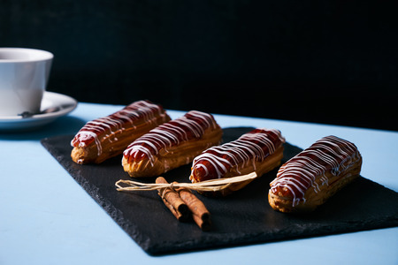 French eclairs with chocolate, tea in cup, tea spoon on saucer and cinnamon sticks on dark slate...