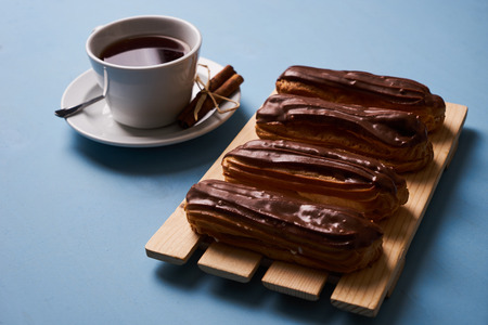 Cup of hot tea with cinnamon steak and french chocolate eclairs on wooden cake board on blue background.の写真素材