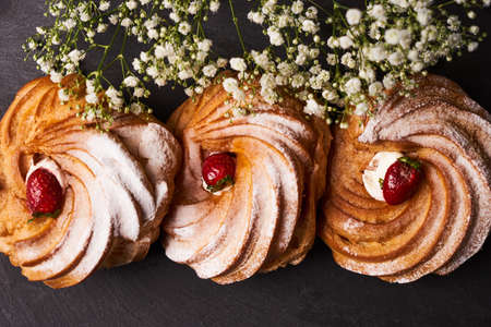 Profiterole, french puff pastry eclairs with custard cream, strawberry and powdered sugar on dark slate cake board on blue background, close-up, top view.の写真素材
