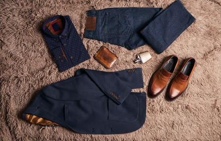 Set of men's clothes such as dark suit jacket with light shirt, jeans trousers, brown leather shoes, belt and wallet on soft carpet background.の写真素材