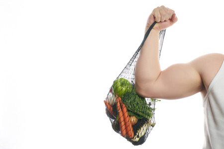 Joyful man athlete holding a shopping bag full of vegetables and flexing his bicep isolated on white background with copy space.の写真素材