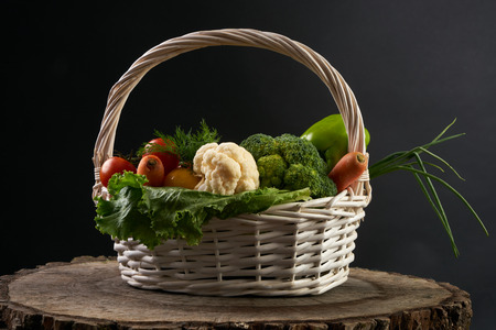 Fresh raw vegetables in a basket. Courgettes, cucumbers, carrots, onions, dill, parsley, broccoli, cauliflower on wooden stump isolated on black background.の写真素材