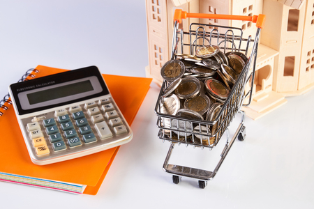 Wood house model, coins in shopping cart and financial notebook on desk table.の写真素材