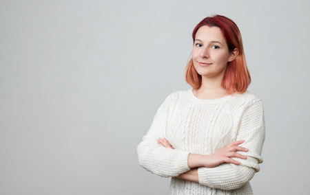 Portrait of young pretty positive girl in a white sweater, smiling looking at camera with crossed arms over white background with copy space.の写真素材