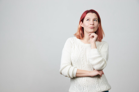 Thoughtful young pretty girl in white sweater holding hand under her head, having doubtful look. Face expressions and emotions.の写真素材