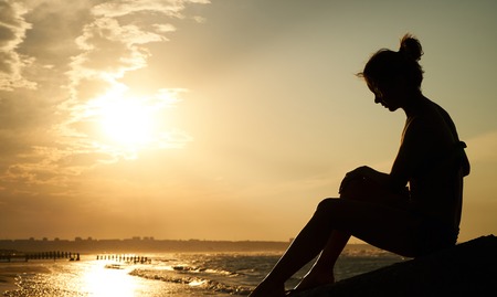 Profile of a sad young woman silhouette in swimsuit, sitting and watching on the beach at sunset. Vacation travel holidays and summer time conceptの写真素材