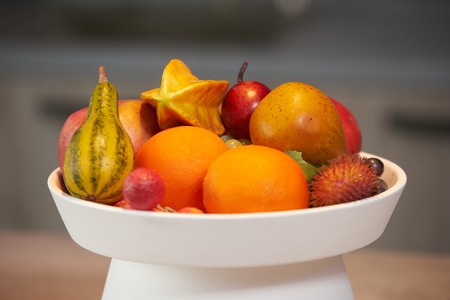 Ripe tropical fruits in a white dish. A plate of tropical fruits and a flower vase standing on the kitchen counter. Luxury modern kitchen interior. Healthy lifestyle and diet food conceptの写真素材