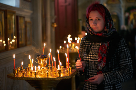 Praying young woman with candle near pedestal with many other candles in church by her hand to show her faith and esteem to Godの写真素材