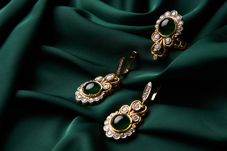 Beautiful Golden ring and pair of earrings with green Emerald and Diamonds gemstones on a green satin background. Luxury female jewelry, close-up. Selective focusの写真素材