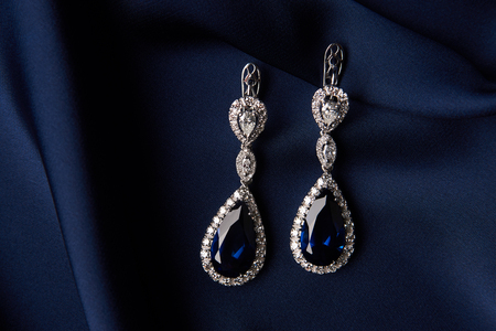 Two Golden sapphire earrings with small diamonds. Pair of platinum earring with sapphire gemstone on blue satin background. Luxury female jewelry, close-upの写真素材