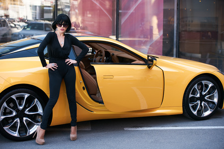 Beautiful brunette woman with yellow sport car. Young woman with dark hair in black bodysuit and sunglasses posing near supercarの写真素材