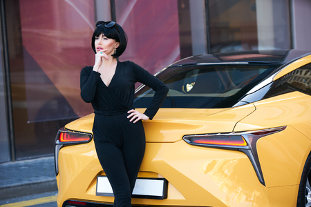 Young woman with dark hair in black bodysuit and sunglasses posing near supercar. Portrait of a Beautiful brunette woman with yellow sport carの写真素材