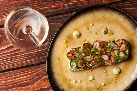 Danish Smorrebrod sandwich with salmon fish, curd mousse, marinated cucumber and radish. Delicious vegetarian snack food with dark rye bread and different topping and wine glass at restaurant tableの写真素材