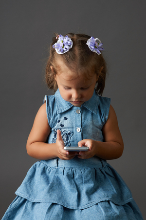 Cute little girl using smartphone on gray background with copy space. Childhood and technology concept. Portrait of little happy girl using cell phoneの写真素材