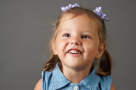 Happy face funny child girl in a blue dress. Portrait of adorable smiling little cute girl child with bow on her head isolated on gray background with copy space.の写真素材