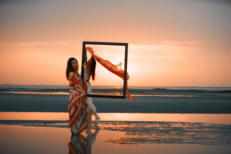 Portrait of a Two Young women holding an empty frame. Girls traveler with wooden picture frame at colorful sea sunset. Summer vacation and travel conceptの写真素材