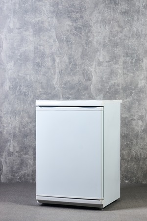 White Empty mini fridge or mini bar against gray textured wall background. International Exhibition furniture elements in large warehouse interior.の写真素材