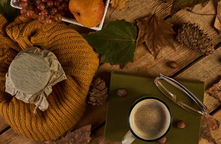 Cozy autumn composition with hot cup of coffee, fresh fruits basket, jam jar, book with reading glasses, nuts and autumn leaves on old wooden table background, top viewの写真素材